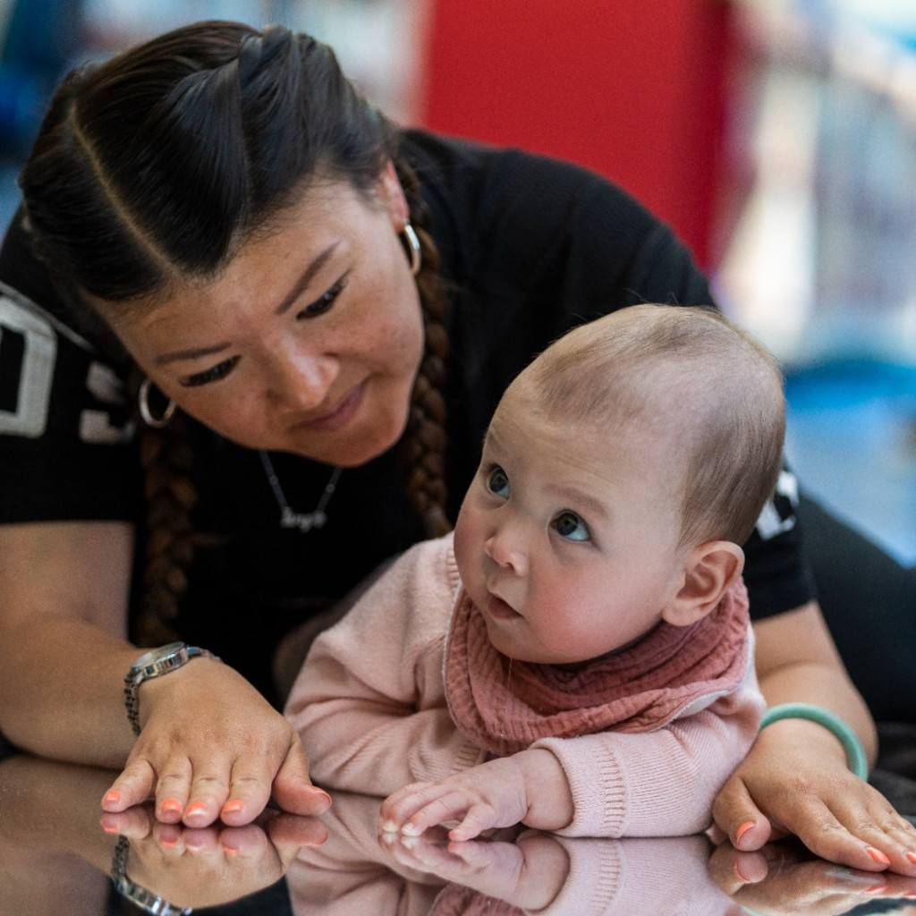 A young child and their carer are lying on their stomachs on top of a reflective, mirrored sheet of card, looking at their reflections.