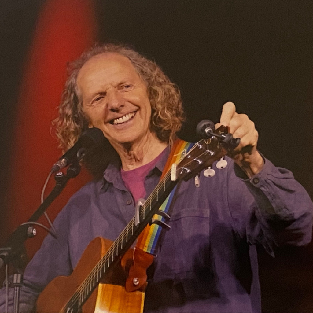 Colum Sands, smiling, standing behind a microphone stand tuning his guitar in front of a red and black background