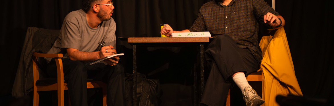 Two men shake hands across a small table during a performance of The Borrowed Room.