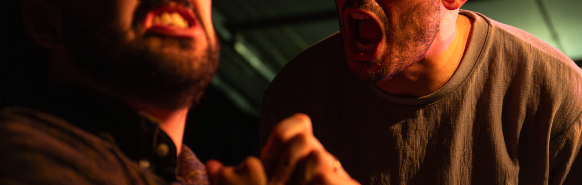Two men shake hands across a small table during a performance of The Borrowed Room.