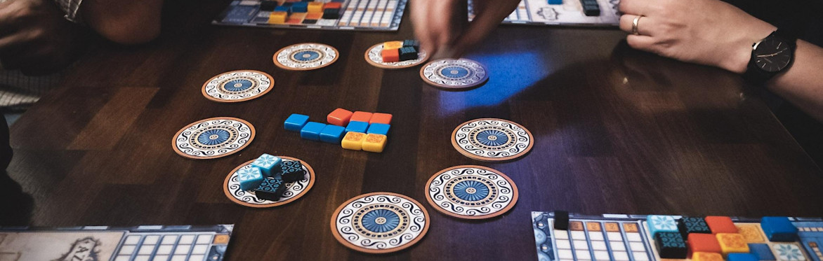 A close up shot of people playing the board game "Azul". There are hands moving to pick up small square colourful tiles from round cardboard tiles. The squares are then placed on each players' board to score points.