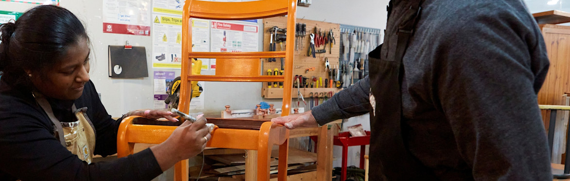 2 people are in a workshop setting, the one on the right is directing the other person how to upholster a dining chair seat with black fabric, using an air powered staple gun.
