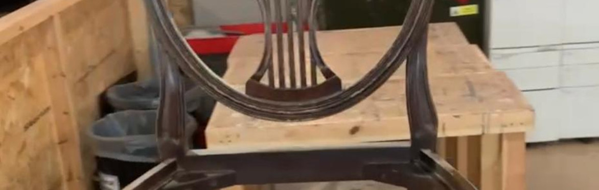 2 people are in a workshop setting, the one on the right is directing the other person how to upholster a dining chair seat with black fabric, using an air powered staple gun.