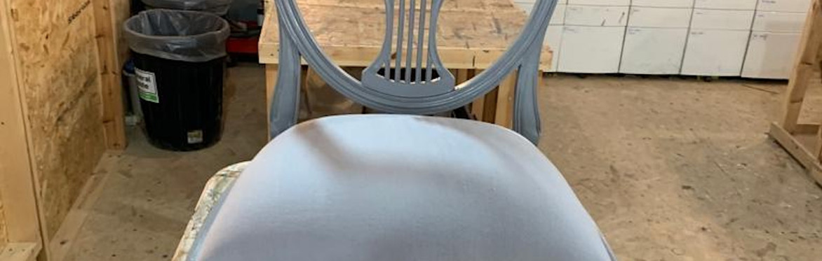 2 people are in a workshop setting, the one on the right is directing the other person how to upholster a dining chair seat with black fabric, using an air powered staple gun.
