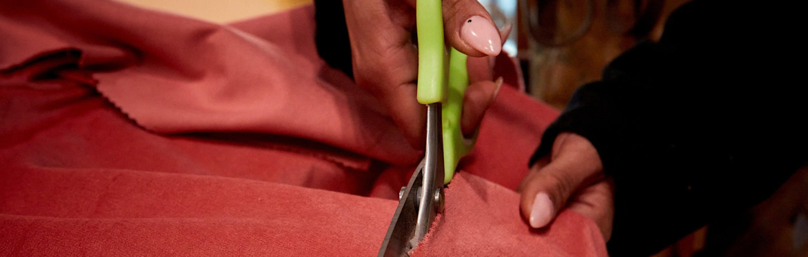 2 people are in a workshop setting, the one on the right is directing the other person how to upholster a dining chair seat with black fabric, using an air powered staple gun.