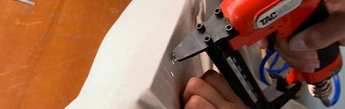 2 people are in a workshop setting, the one on the right is directing the other person how to upholster a dining chair seat with black fabric, using an air powered staple gun.