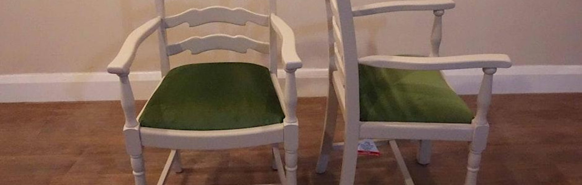 2 people are in a workshop setting, the one on the right is directing the other person how to upholster a dining chair seat with black fabric, using an air powered staple gun.