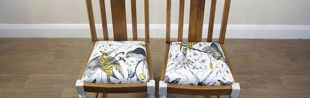 2 people are in a workshop setting, the one on the right is directing the other person how to upholster a dining chair seat with black fabric, using an air powered staple gun.