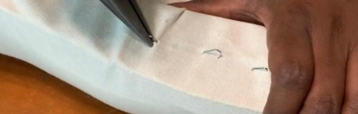2 people are in a workshop setting, the one on the right is directing the other person how to upholster a dining chair seat with black fabric, using an air powered staple gun.