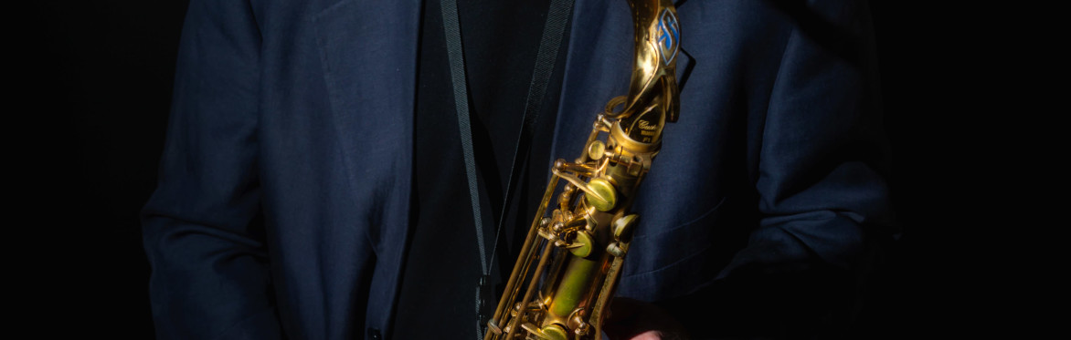 Saxophonist stands holding a tenor saxophone under a spotlight, wearing a dark jacket against a black background. The close portrait captures a calm, reflective expression.