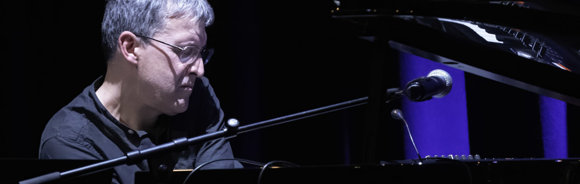 Pianist performs at a grand piano under blue stage lighting. A microphone is positioned above the instrument as he concentrates on playing against a dark concert backdrop.