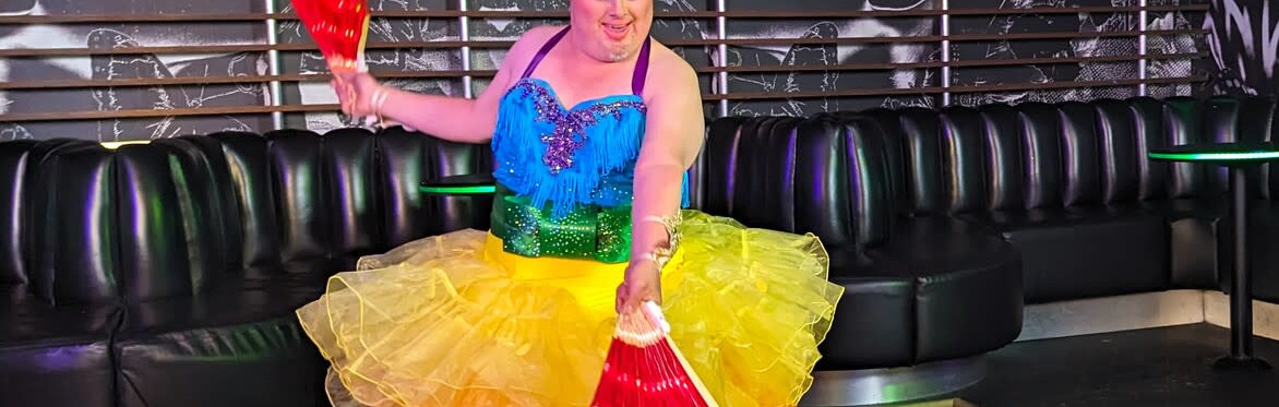 Performer in colourful drag costume poses in a nightclub, wearing a sparkling headdress with bright rainbow feathers, dramatic blue eye make-up, gold gloves and a vivid green and blue outfit.