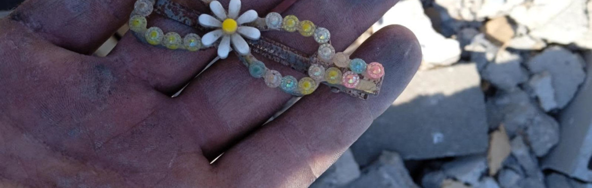 A hand holds a child's hair clip with a flower on it in front of rubble.