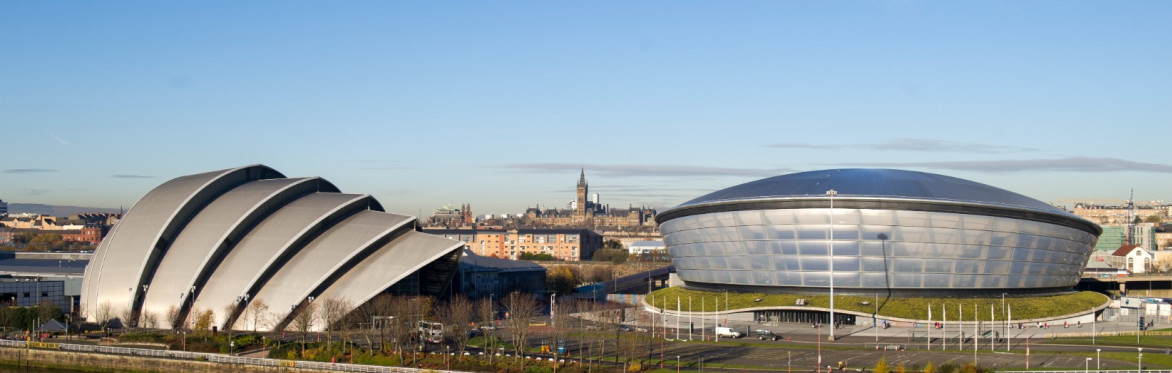 Panoramic view of Glasgow's waterfront showing the SEC Armadillo and OVO Hydro beside the River Clyde, with reflections in the water and the city skyline in the distance.