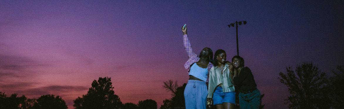 Three Black people stand in the outside smiling against a bright night sky.