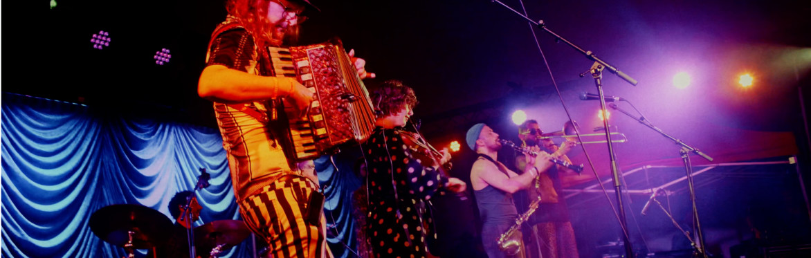 A drummer, accordionist, violinist, clarinetist and trombonist are in a colourfully lit festival stage playing their instruments
