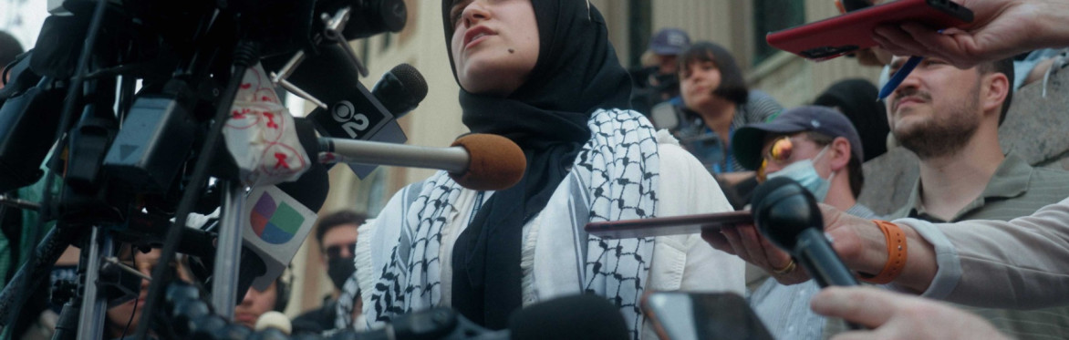 Young woman wearing a hijab and keffiyeh speaking into news companies' microphones outside Columbia University