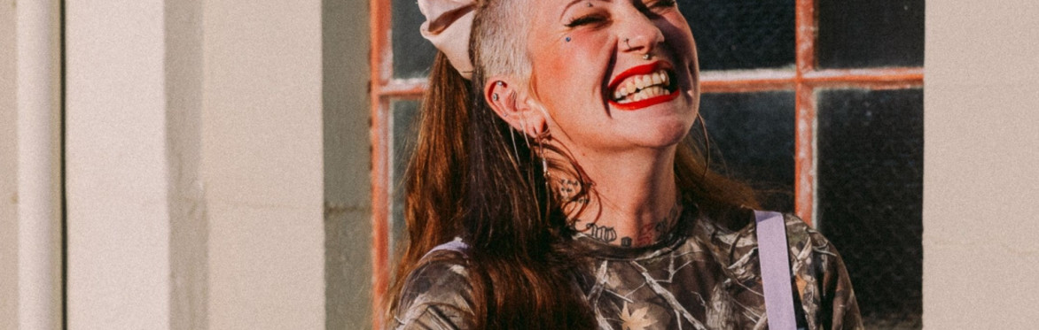 Colour photograph of folk singer Jennifer Reid. Her hair is shaved at the sides with a long, red ponytail at the back. Jennifer is wearing red lipstick and laughing.