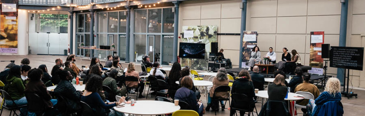 A gathering of people clustered around small tables listening to speakers on the stage to the right of the image