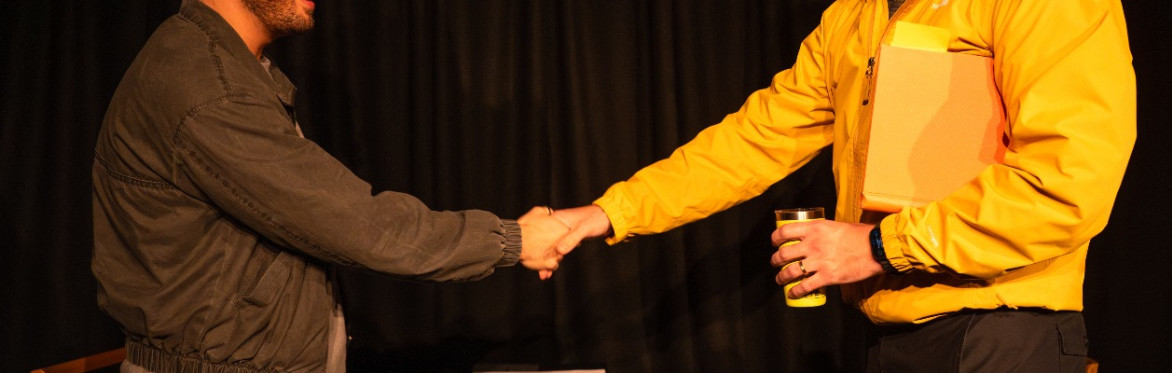 Two men shake hands across a small table during a performance of The Borrowed Room.