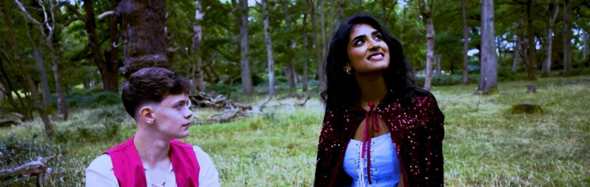 Picture of a woman dressed in a fairytale outfit with a sparkling red cape together with a  young man dressed as a fairytale hero. They are sitting within woodland.