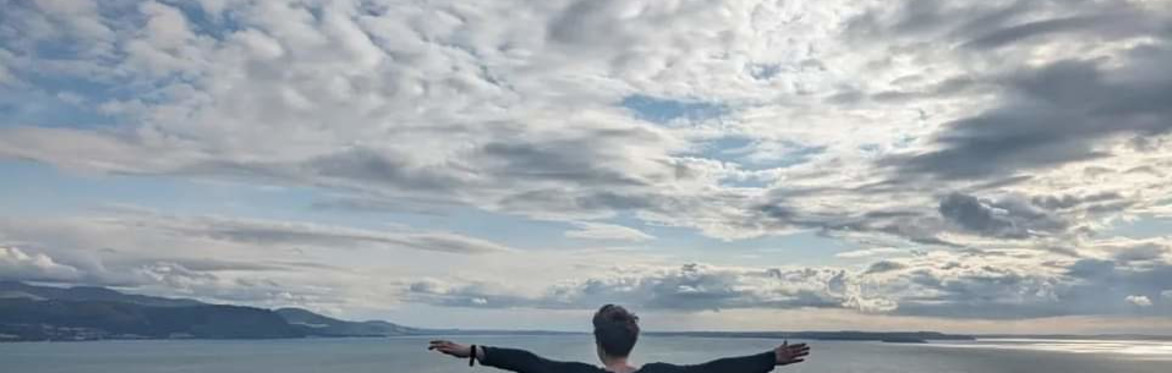 Becky, a white woman in her 50s with short hair from the back stands, arms stretched out almost in line with the horizon above the sea. The sky is blue with dramatic white clouds.