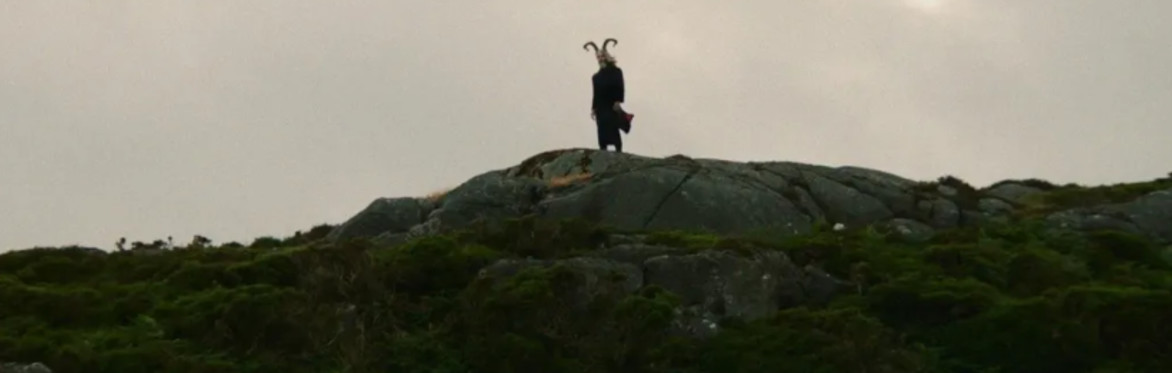 A man with horns and a black coat stands on a rocky outcropping