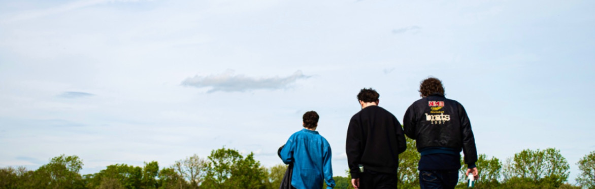 The three band members walking away surrounded by trees and a sunny sky