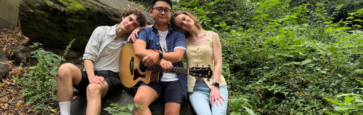white man with a white shirt sitting on a rock leaning on his asian male friend, who sits in the middle with a guitar. To his right leaning on him too, a white woman with a yellow sleeveless top.