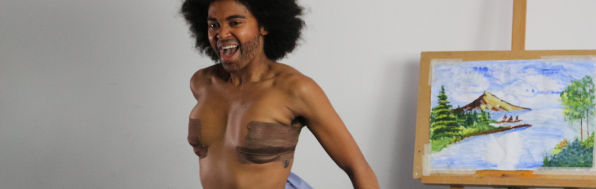 A Black drag king stands topless next to a canvas painting.