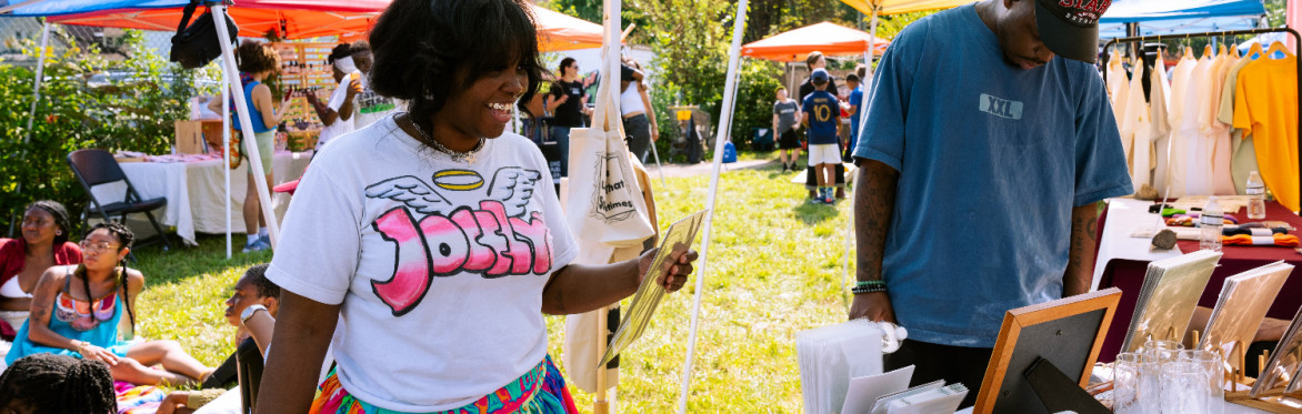 A sunny outdoor festival with two Black people interacting with a vendor