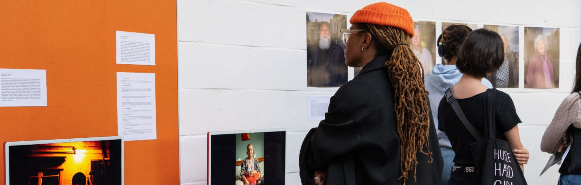 Person with a black coat and orange beanie hat, looking at exhibition submissions on a white wall