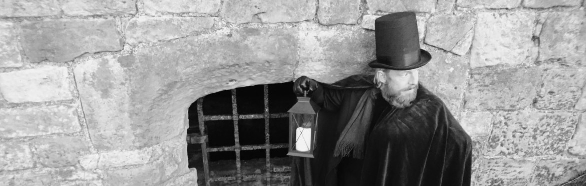 A performer from Time Will Tell Theatre Company. stands with a lantern outside a metal grate in a stone wall
