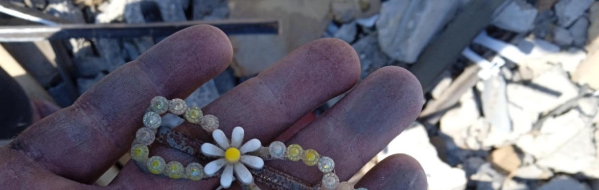 A hand holds a child's hair clip with a flower on it in front of rubble.
