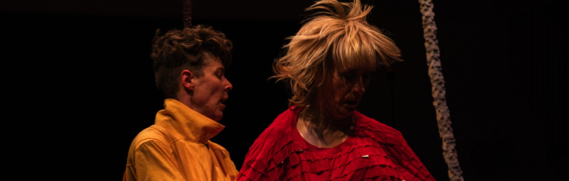 A blond-haired person in a red shirt lifts upward while another person in yellow holds them in a partial embrace.