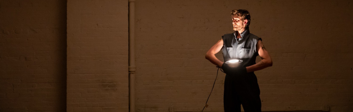 A performer stands against a large brick wall in a dim, warm-toned industrial space. Wearing a dark sleeveless outfit, they hold a glowing circular light at their waist, illuminating their face and to