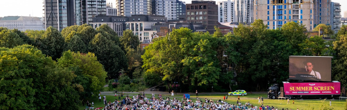 A large crowd sits on the grass in a park, watching a film on a large screen.