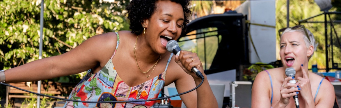 Two women sing into microphones at an outdoor karaoke session in a park.