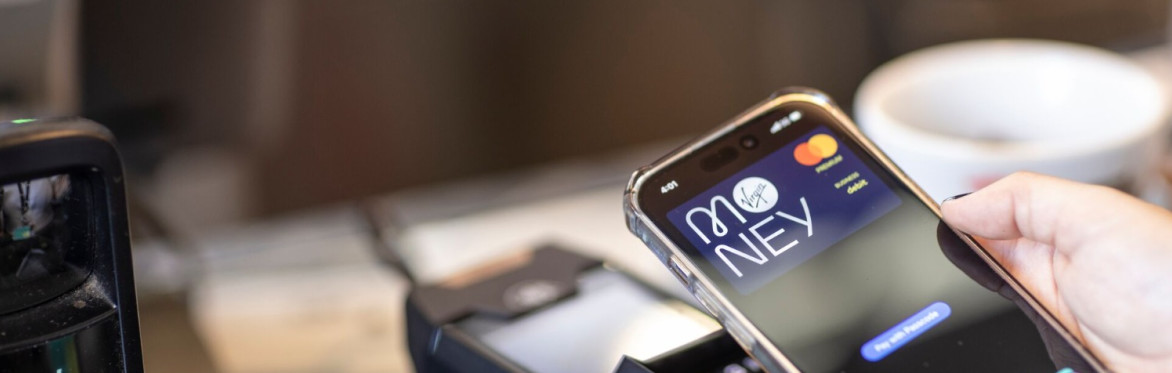 Close-up of a mobile phone displaying a Virgin Money payment screen held above a card reader on a glass counter indoors. A white cup and part of a barcode scanner are visible in the background.