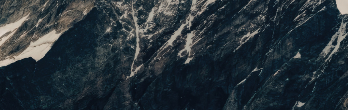 Snow-dusted rugged mountain peak under a cloudy sky, with dramatic dark rock formations. The scene conveys a sense of solitude and awe.