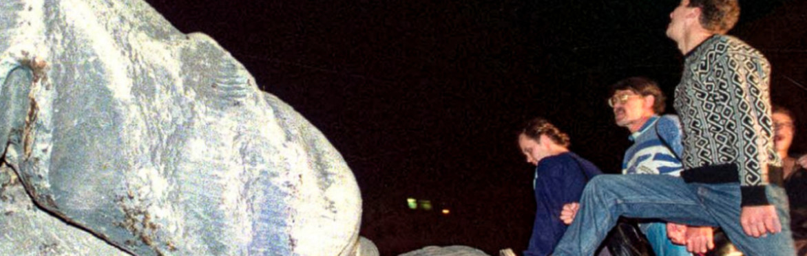 A group of people walks across a toppled statue of Lenin at night, one man stepping on its head. The scene conveys defiance and change.