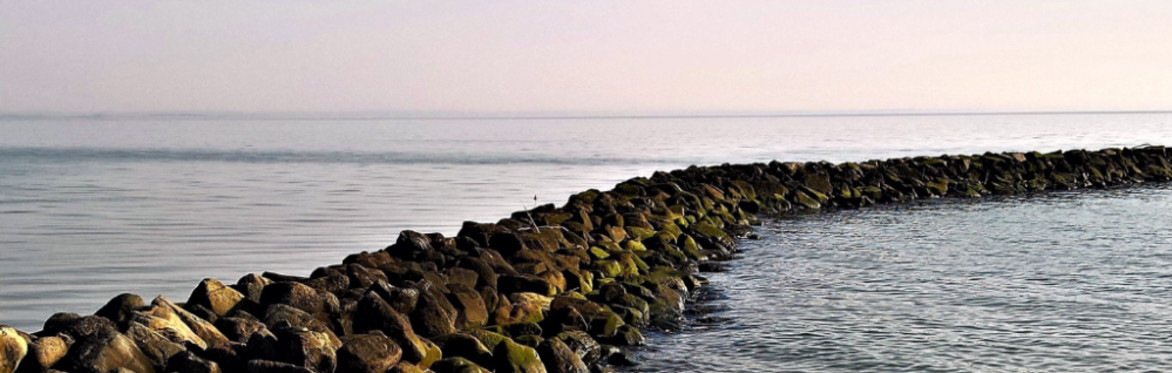 Curved rock barrier extends into calm sea under a blue sky, creating a serene, peaceful coastal scene. Cooper Education logo appears on the top right hand side.