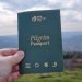 a hand holds a green pilgrim passport with a hillside in the background