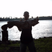 A man in a hat backlit by morning light standing on the shore with a river in the background holding a large catfish