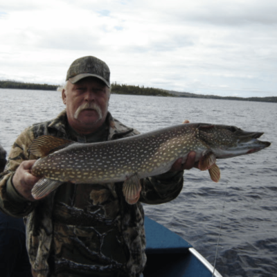 A man with a gray mustache, camouflage hat and jacket, on a lake in a boat holding a northern pike