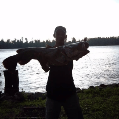 A man in a hat backlit by morning light standing on the shore with a river in the background holding a large catfish