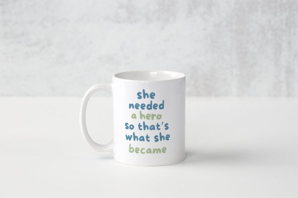 A white ceramic mug with a light blue and green text saying 'she needed a hero so that's what she became'.