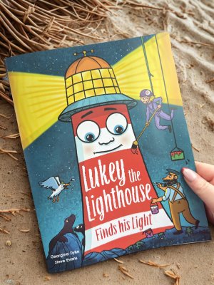 Image shows a copy of 'Lukey the Lighthouse finds his Light' book on a sandy beach.