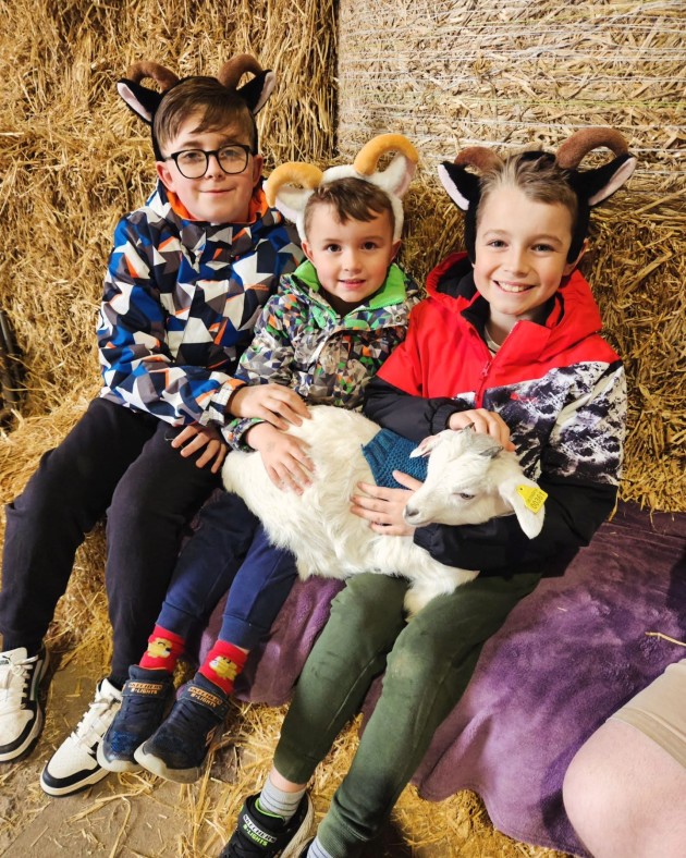 Three young boys smiling wearing goat horns and ears sitting on hay bales cuddling a baby goat wearing a knitted jumper