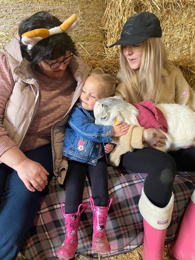 A granny, mother and child smiling sitting on hay bales whilst cuddling a baby goat